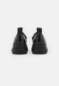 Czarne skórzane sneakersy z grubą podeszwą, złotymi akcentami, pętlami do wkładania oraz wytłoczonym logo na pięcie.