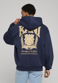 Felpa con cappuccio blu navy caratterizzata da un grande logo beige e testo sulla parte posteriore con dettagli grafici. Tessuto morbido e design dalla vestibilità rilassata.