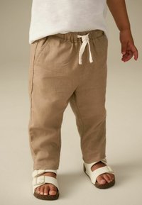 Pantaloni in lino marrone chiaro con cintura elastica e lacci. Presentano tasche laterali e una vestibilità comoda, abbinati a sandali bianchi.