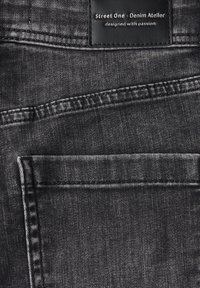 Tessuto di denim nero con una texture liscia. Presenta un'etichetta con la scritta "Street One - Denim Atelier" e una tasca posteriore. Cuciture visibili.
