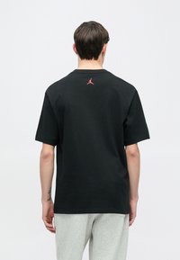 Μαύρο βαμβακε ρό μπλουζάκι με κοντά μανίκια, με μικρό κόκκινο λογότυπο Jordan στο πίσω μέρος του λαιμού. Φορεμένο με ανοιχτό γκρι παντελόνι.