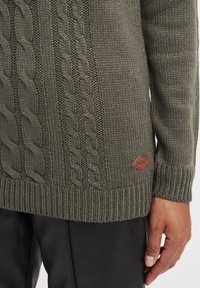 Pull en tricot vert olive avec des motifs en torsade, ourlet et poignets côtelés, et une étiquette en cuir marron sur le côté.