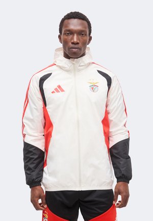 Muž tvárou vpredu, nosiaci bielo-červeno-čiernu športovú bundu na zips s logom Adidas a emblémom futbalového klubu Benfica.