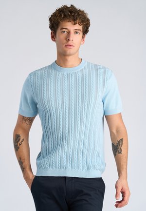 Joven con cabello rizado que lleva un suéter de punto de manga corta azul claro y pantalones oscuros, mostrando tatuajes de mariposas y flores en los brazos.