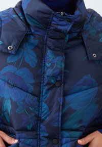 Giacca a vento navy con motivo floreale blu. Caratterizzata da un colletto alto, chiusure a pressione e texture trapuntata. Tessuto morbido e leggero.