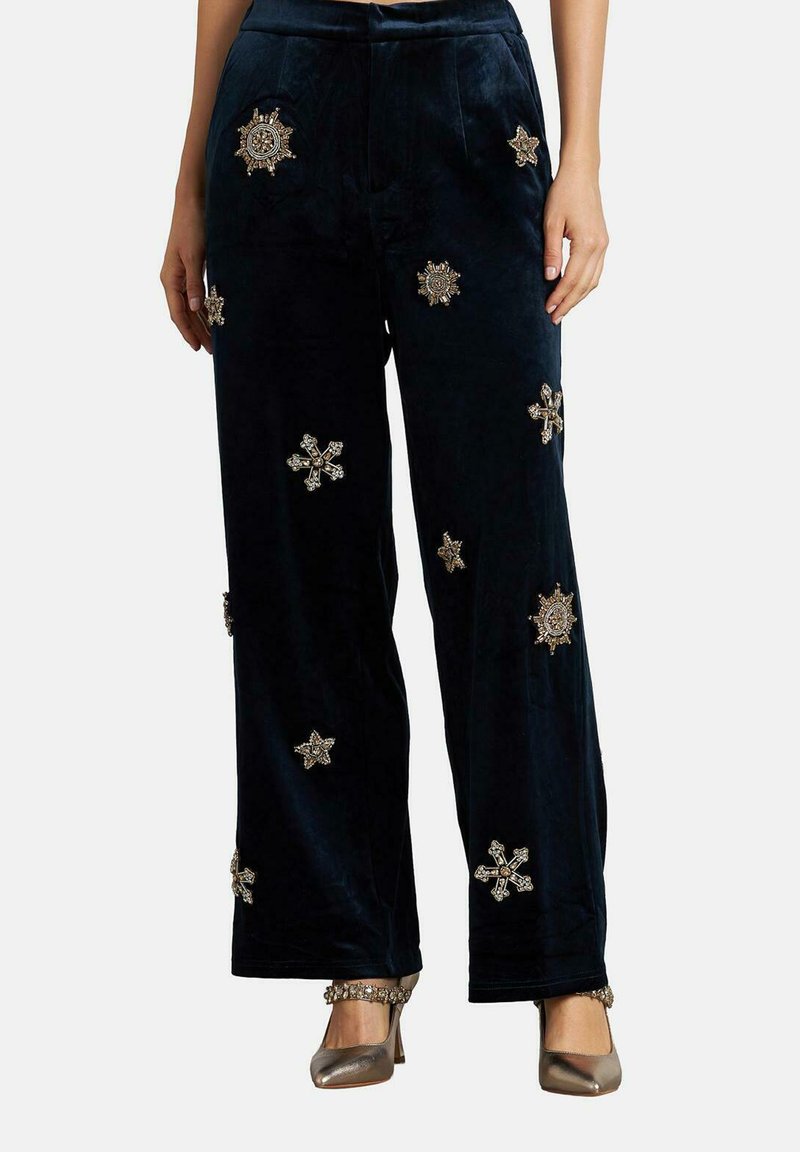 Pantalones de terciopelo azul marino con corte de pierna ancha, adornados con patrones de estrellas y copos de nieve en tono dorado, con una textura suave y un corte a la altura del tobillo.