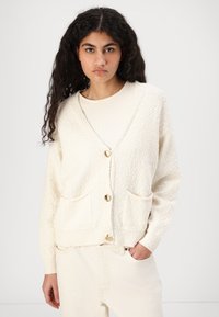 Creme strikket cardigan med et struktureret mønster, v-hals, to frontlommer og guldknapper. Bæres over en lys top og kombineres med jeans.