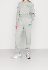 Kvinna iklädd en grå Nike-tröja och sweatpants med gröna logotyper, kombinerat med pastellfärgade lila och orange sneakers, stående mot en vit bakgrund.