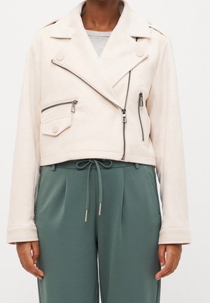 Femme portant une veste courte écrue avec des revers et des fermetures éclair visibles sur une chemise gris clair et un pantalon vert à cordon.