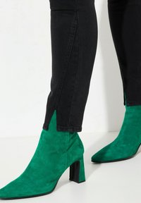 Botines de ante verde con punta afilada y tacón cuadrado, combinados con jeans negros ajustados con slit en los laterales.