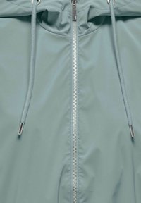 Veste à capuche vert clair avec fermeture éclair frontale visible et cordons avec embouts métalliques.