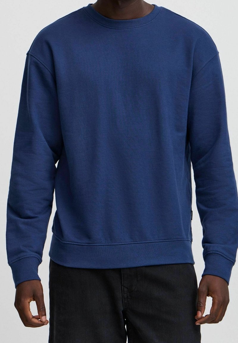 Homme portant un sweat-shirt bleu uni à manches longues et un pantalon noir, debout devant un fond gris clair.