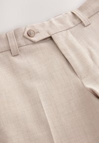 Gros plan sur la ceinture d’un pantalon en lin beige avec fermeture par bouton, passants pour ceinture et tissu lisse légèrement texturé.