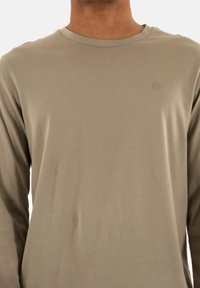 Camicia a maniche lunghe in tessuto beige chiaro, con scollo rotondo e un logo sottile dello stesso colore sul petto. Texture liscia.