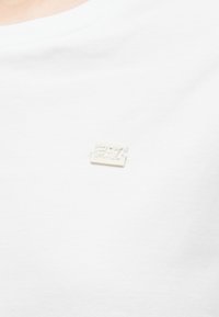 Biele bavlnené tričko s malou obdĺžnikovou kovovou značkou s vyrazeným textom na ľavej strane hrudníka.