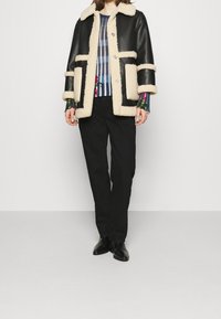 Svart läderjacka med beige foder i fleece, med rund krage, dubbla sidofickor och knappstängning. Randig skjorta och svarta byxor bärs under.