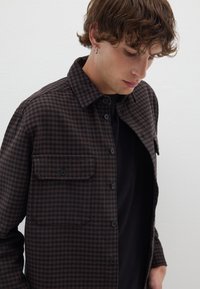 Bruine en zwarte getailleerde overhemdjacket met een knoopsluiting aan de voorkant, twee borstzakken en een textuurstof. Draagt over een zwart shirt.
