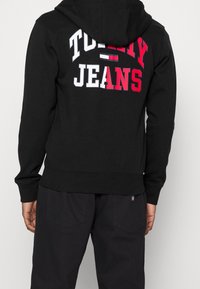 Felpa nera in cotone, con la scritta "TOMMY" e "JEANS" in lettere rosse e bianche sul retro. Include una tasca a marsupio e polsini a costine.