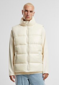 Puffer vest in cream, with a high collar, zip closure, and horizontal quilted pattern. Worn over a long-sleeve shirt, paired with light jeans.