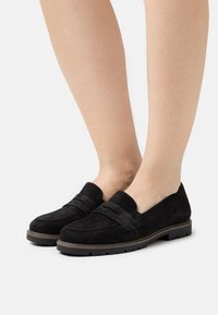 Mustat mokkanahkaiset loaferit, joissa on sileä pinta, leveällä nauhalla ja matala kumipohja, nähtynä sivulta, paljain säärin näkyvissä.