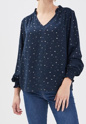 Blouse bleu marine avec un col en V et un col froncé. Présente des motifs floraux et des pois argentés, ainsi que des poignets smockés. Tissu léger.