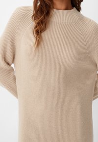 Beige pletený svetr s vysokým límcem, žebrovanou texturou a raglánovými rukávy, s volným střihem a měkkým materiálem.