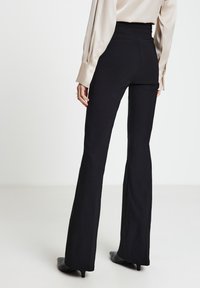 Schwarze ausgestellte Hose mit hoher Taille aus glattem Stoff. Rückennahtdetails und eine dezente Textur, kombiniert mit einer hellfarbenen Bluse.