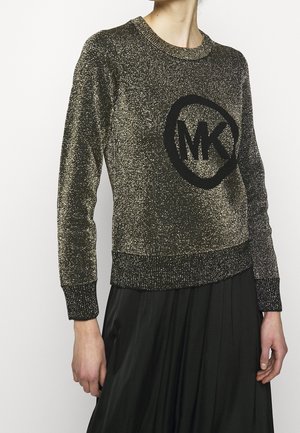 Femme portant un pull à manches longues doré scintillant avec un logo "MK" noir et une jupe plissée noire, debout devant un fond uni.