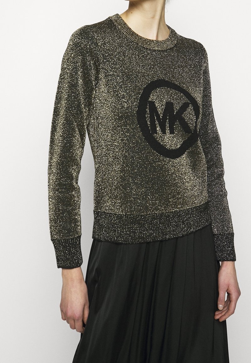 Femme portant un pull à manches longues doré scintillant avec un logo "MK" noir et une jupe plissée noire, debout devant un fond uni.