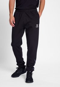 Pantalones de chándal negros con cintura elástica, bolsillos laterales y logo. Tiras laterales en gris. Tejido suave y casual. Combinados con zapatillas deportivas negras.