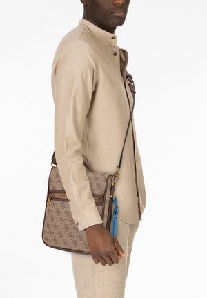Borsa a tracolla con motivo marrone, ferrature dorate, tasche con cerniera e un pendaglio blu. Indossata da una persona con una camicia beige chiaro.