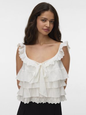 Jonge vrouw met donker golvend haar die een witte mouwloze top draagt met gerafelde lagen en een strik aan de voorkant, staand tegen een effen achtergrond.