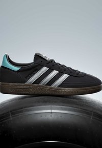 Baskets Adidas noires avec des bandes argentées, semelle en gomme, languette de talon bleu sarcelle, présentées sur un grand pneu noir sur fond uni.