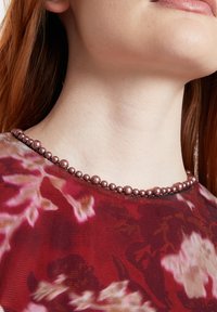 Rote Bluse mit floralem Muster, die eine transparente Textur aufweist und mit einem mit Perlen verzierten Ausschnitt in Rosatönen und metallischen Akzenten versehen ist.