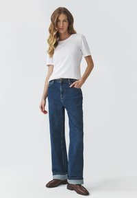 Camiseta blanca de manga corta combinada con jeans de pierna ancha de tiro alto en azul oscuro con dobladillos rematados, acentuada con un cinturón marrón y zapatos de cuero.