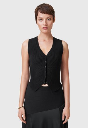 Kvinne med kort brunt hår iført en svart ermeløs ribbestrikket vest med knapper og svart skjørt, stående foran en grå bakgrunn.