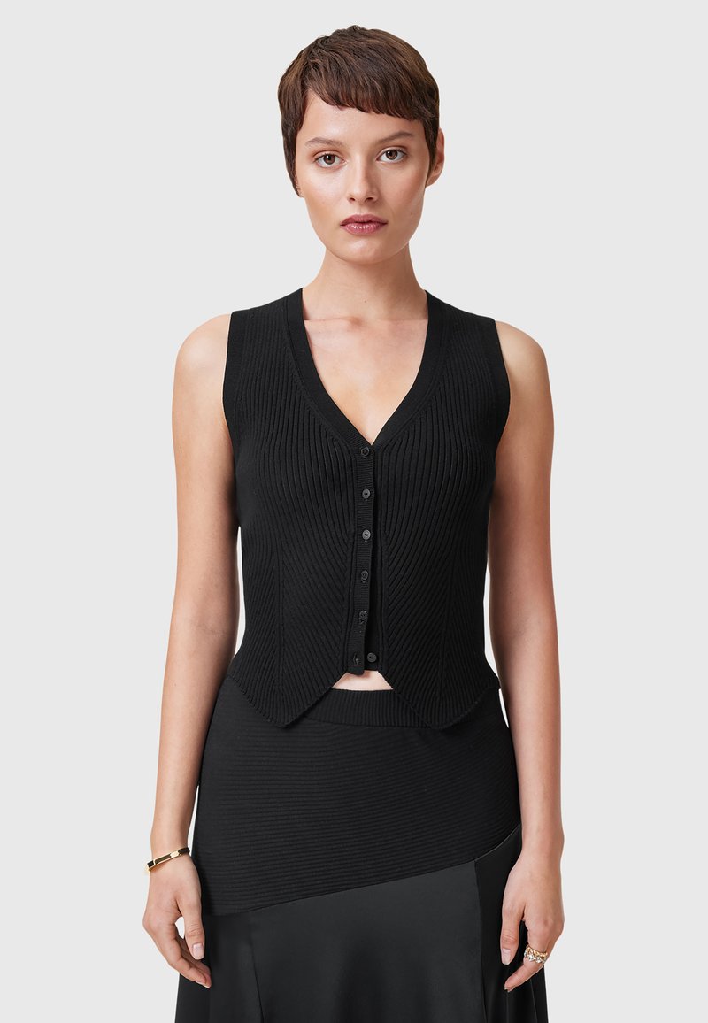 Kvinde med kort brunt hår iført en sort ribbet vest uden ærmer med knapper og sort nederdel, stående mod en grå baggrund.