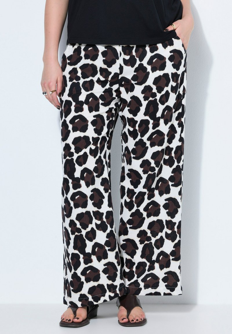 Bukser med brede ben med sort og brun leopardprint, båret med sort top og sorte sandaler med åbne tæer, hænder i lommerne og ringe på fingrene.