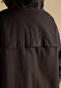 Tumši brūns kapuce jaka ar horizontālu aizmugures atloku detaļu un smalku "JOULES" zīmolu augšējā aizmugurē.