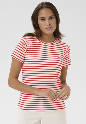 Žena s hnedými vlasmi, oblečená v bielom tričku s červeno-bielymi pruhmi s krátkym rukávom a svetlých nohaviciach, stojaca s jemne zloženými rukami.