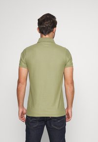 Homme portant un polo à manches courtes vert olive et un jean foncé, tourné dos à un fond clair.