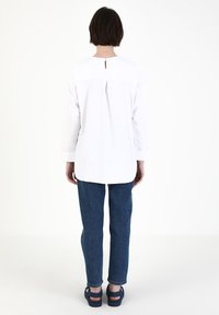 Hvid langærmet skjorte med en nøglehul-detail på ryggen, parret med blå denimbukser og sandaler. Glat tekstur, afslappet pasform og lige kant.