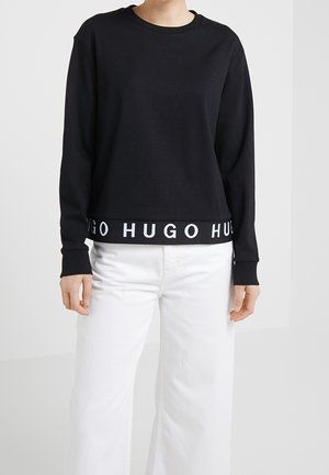 Persoon met een zwart sweatshirt met tekst "HUGO" op de zoom en een witte broek, staand tegen een effen lichte achtergrond.