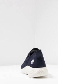 Chaussure de sport bleu marine avec une tige tricotée texturée, une semelle en caoutchouc blanche, des lacets élastiques et un logo sur le talon. Tirette arrière lisse pour un enfilage facile.