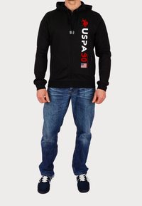 Felpa con cappuccio nera con zip, stampa rossa "USPA 90", cappuccio e laccetti. Indossata con jeans blu e sneakers scure. Dettaglio della bandiera americana visibile.
