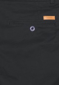 Zadní kapsa černých kalhot s knoflíkovým zapínáním, na které je vytištěn text "ESPRIT", a malá kožená značka hnědé barvy přišita nad kapsou.