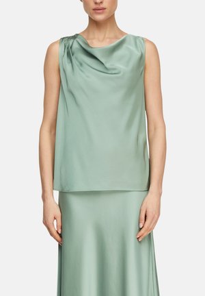 Kobieta ubrana w bezrękawnikowy, szałwiowozielony satynowy top z dekoltem z marszczeniem oraz dopasowaną spódnicę, stojąca z rękami swobodnie opuszczonymi wzdłuż ciała.