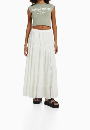Cropped t-shirt verde con scritte bianche, abbinato a una lunga gonna bianca a balze. Sandali neri con plateau completano l'outfit.