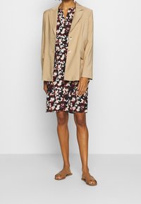 Blazer beige con dos botones y bolsillos con solapa sobre un vestido negro con estampado floral. La modelo lleva sandalias planas marrones.