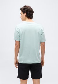 Άνδρας που στέκετα ι με την πλάτη, φοράει ανοιχτό μπλε κοντομάνικο πουκάμισο και μαύρο σορτς, σε φόντο απλώς λευκό.